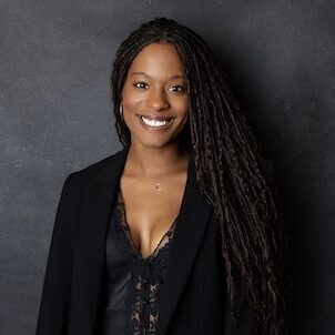 A professional portrait of a smiling woman with long locs wearing a black blazer over a lace top. She is posed against a dark, textured studio background.