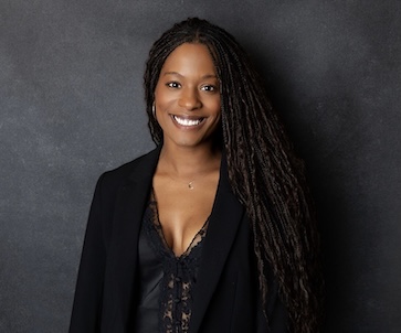 A professional portrait of a smiling woman with long locs wearing a black blazer over a lace top. She is posed against a dark, textured studio background.