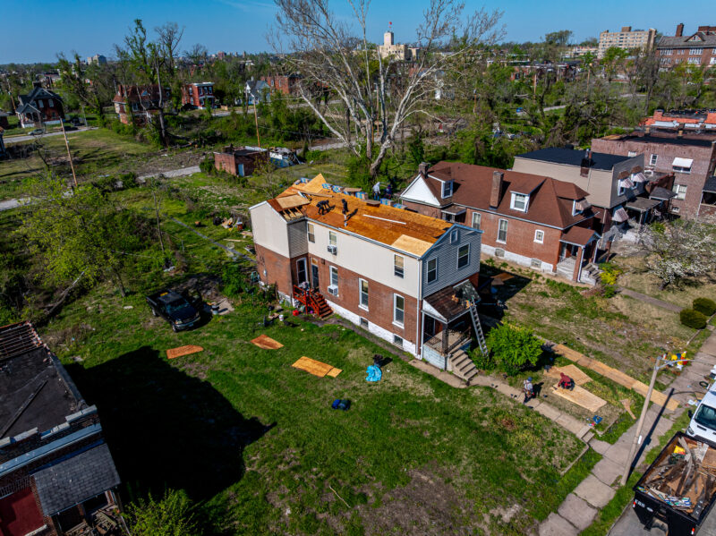 A high-angle aerial view shows a house undergoing a major roof replacement within a neighborhood that shows signs of prior storm damage, including damaged trees and neighboring roofs. A construction dumpster sits on the street in the lower right corner.
