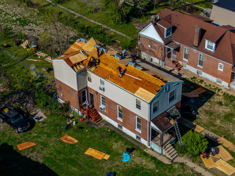 A wide drone photo captures a team of workers replacing the roof on a two-story brick and siding house in a residential neighborhood. Construction materials and a parked truck are visible in the grassy yard below.