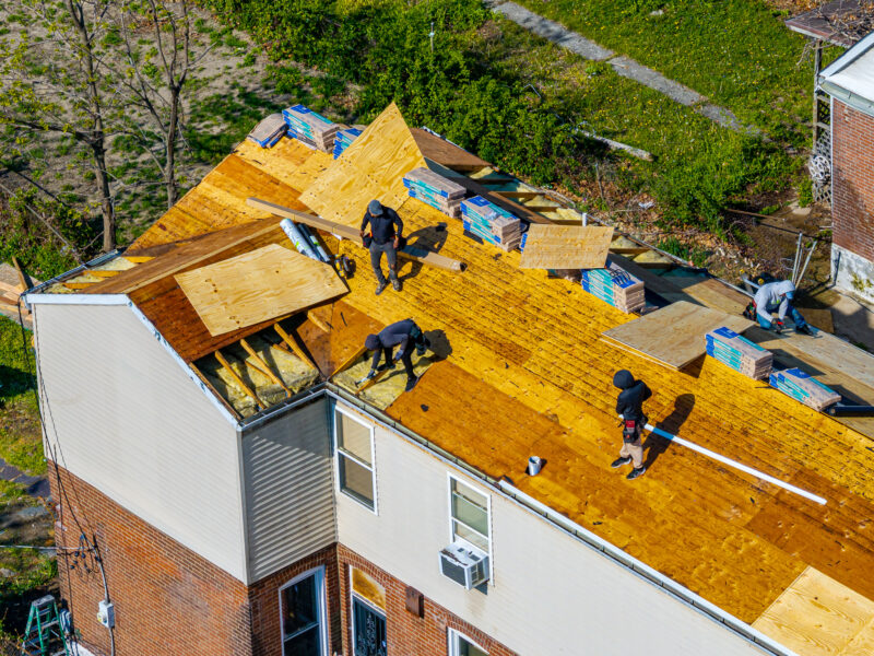 An aerial shot shows several construction workers on the roof of a two-story house installing new wooden plywood sheathing. Stacks of shingles and roofing materials are staged across the exposed roof trusses.
