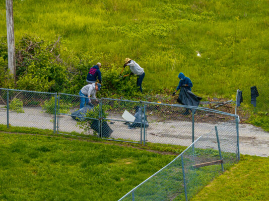 A group of four volunteers uses shears to clear overgrown weeds and brush along a chain-link fence bordering a grassy field. They are bagging the yard waste into large black trash bags.