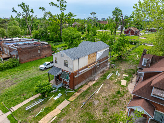 An elevated view shows a two-story house undergoing exterior repairs, with scaffolding erected along a side wall that has missing siding and exposed wooden panels. Construction materials and debris are scattered across the grassy yard surrounding the home.