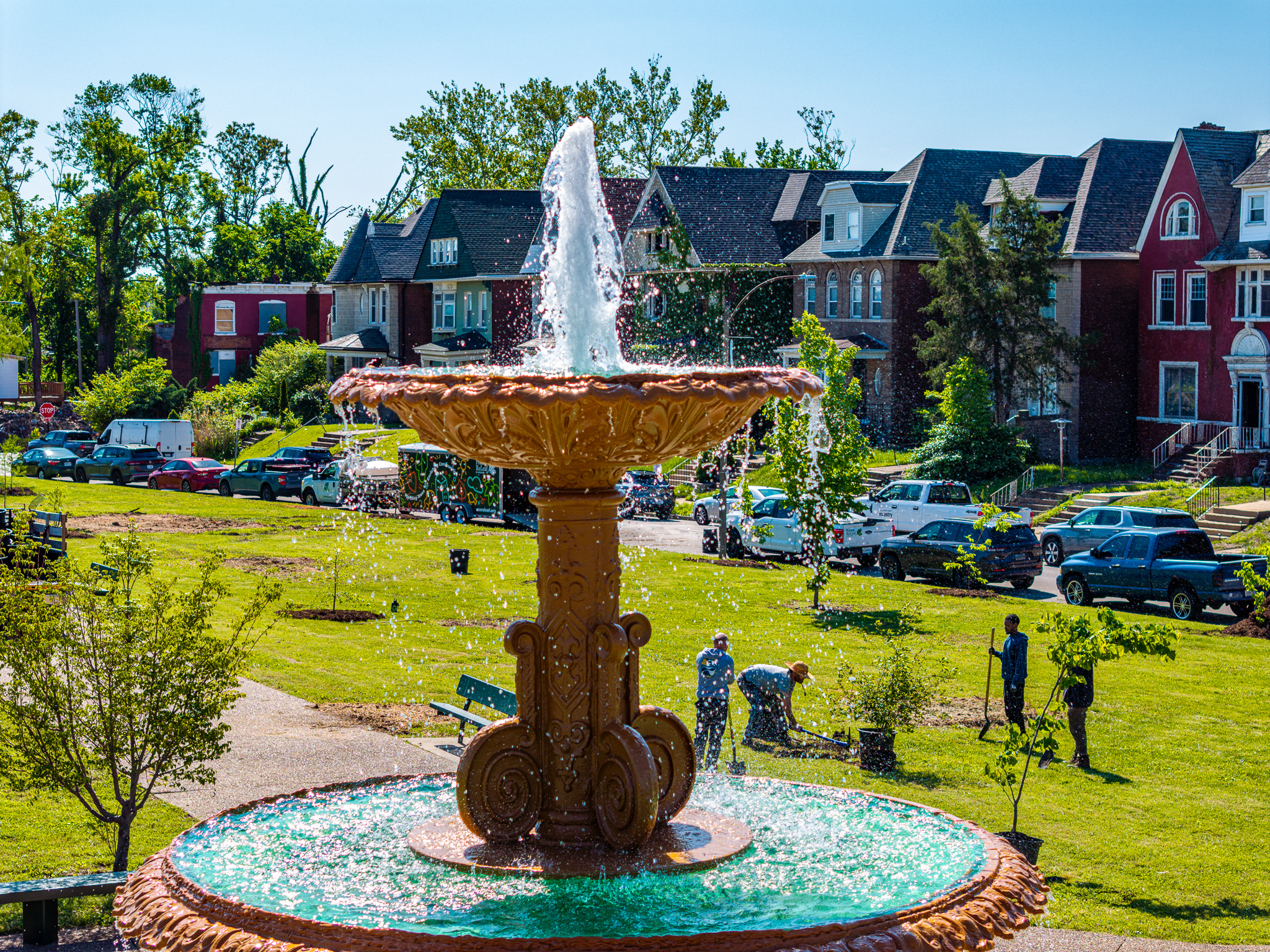 An ornate, multi-tiered fountain sprays sparkling water in the foreground of a sunny park. In the background, volunteers are actively planting young trees in the grass across from a row of historic brick houses.