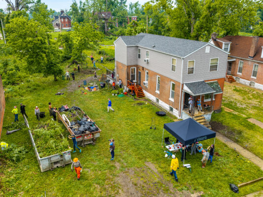 An aerial view shows volunteers working in an empty grassy lot behind a brick building, with a black canopy tent set up on the lawn. Several people are loading freshly cut tree branches and brush into a large open dumpster.