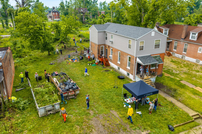 An aerial view shows volunteers working in an empty grassy lot behind a brick building, with a black canopy tent set up on the lawn. Several people are loading freshly cut tree branches and brush into a large open dumpster.