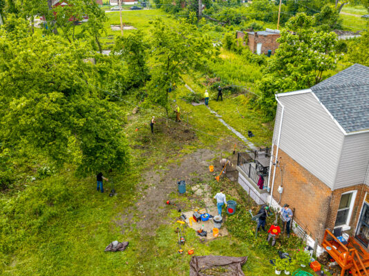 An aerial shot captures a team of volunteers clearing brush and cutting down a large tree in the messy backyard of a two-story brick house. Workers are scattered across the yard using various tools to process the downed branches.