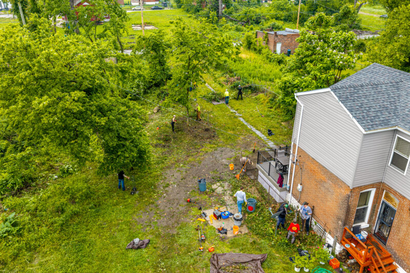 An aerial shot captures a team of volunteers clearing brush and cutting down a large tree in the messy backyard of a two-story brick house. Workers are scattered across the yard using various tools to process the downed branches.