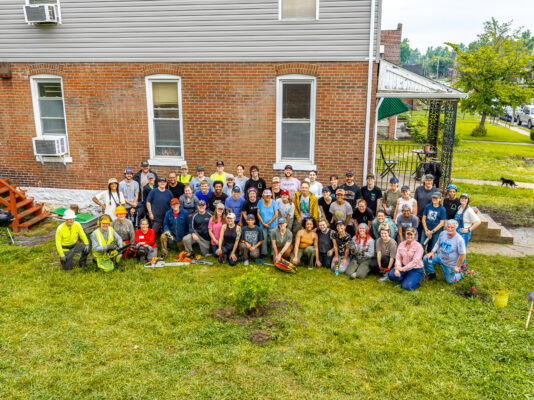A large group of smiling volunteers poses for a photo in the backyard of a brick building, holding various landscaping tools like chainsaws, shovels, and rakes. The diverse group is wearing casual work clothes, with some sporting safety helmets and high-visibility vests.
