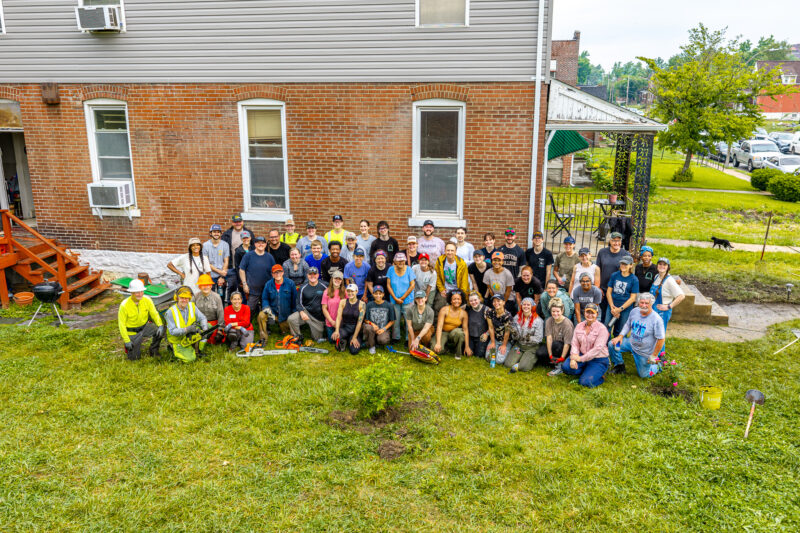 A large group of smiling volunteers poses for a photo in the backyard of a brick building, holding various landscaping tools like chainsaws, shovels, and rakes. The diverse group is wearing casual work clothes, with some sporting safety helmets and high-visibility vests.