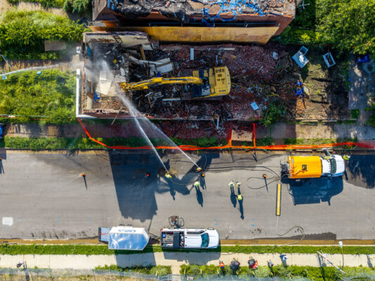A top-down aerial view reveals a yellow excavator parked directly on top of a demolished building's rubble. Below, workers use hoses connected to a yellow street-parked truck to spray down the dusty site.