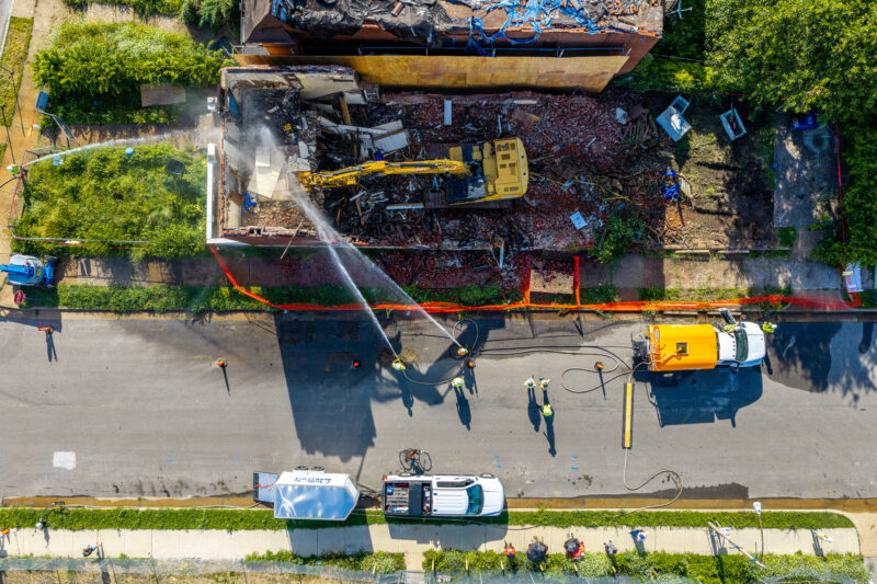 A top-down aerial view reveals a yellow excavator parked directly on top of a demolished building's rubble. Below, workers use hoses connected to a yellow street-parked truck to spray down the dusty site.