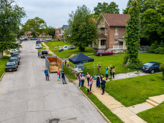 An elevated view of a residential street shows volunteers gathered around a black "Grassroots" canopy tent and a large, open debris dumpster.