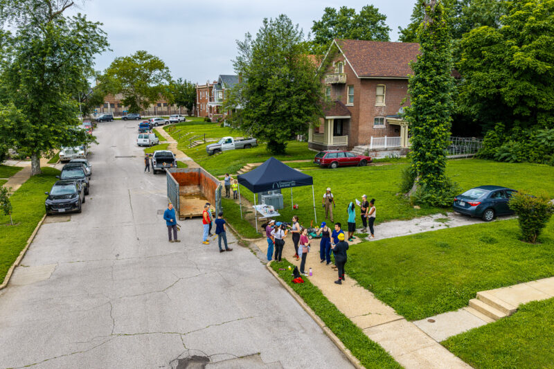 An elevated view of a residential street shows volunteers gathered around a black "Grassroots" canopy tent and a large, open debris dumpster.