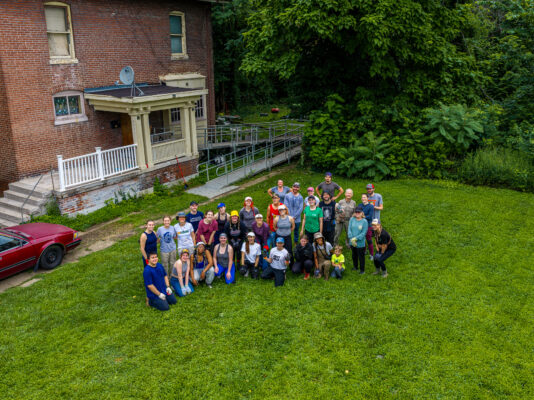 An aerial drone photo features a large group of smiling volunteers waving at the camera from the grassy lawn of a multi-story brick house with a wheelchair ramp.