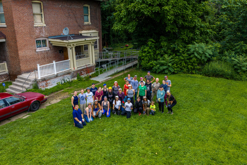 An aerial drone photo features a large group of smiling volunteers waving at the camera from the grassy lawn of a multi-story brick house with a wheelchair ramp.