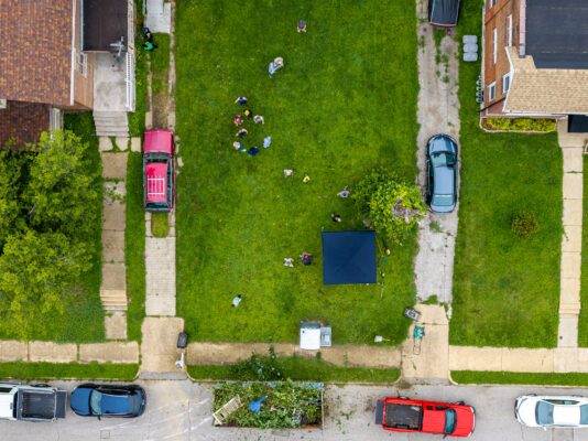 A top-down aerial shot captures a group of people scattered across a large green lawn doing yard work. A dark canopy tent, parked cars, and a yard waste dumpster sit at the edges of the property.