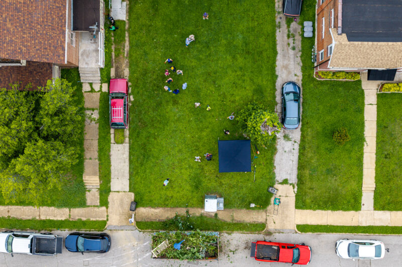 A top-down aerial shot captures a group of people scattered across a large green lawn doing yard work. A dark canopy tent, parked cars, and a yard waste dumpster sit at the edges of the property.