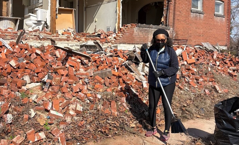 A woman wearing a face mask and dark vest holds a broom while standing in front of a massive pile of red bricks from a collapsed building. The exposed interior of the ruined structure is visible directly behind her.