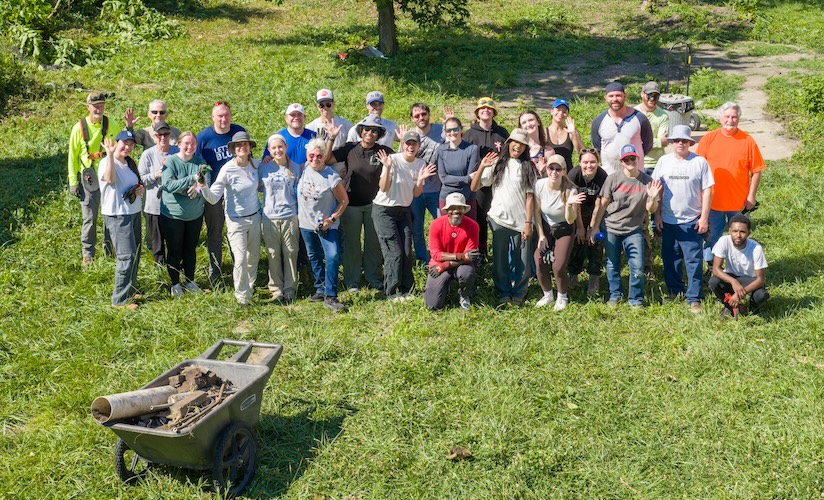 A group of over twenty smiling volunteers waves at the camera while standing in a sunlit grassy field. In the foreground, a wheelbarrow is loaded with discarded bricks and construction debris.