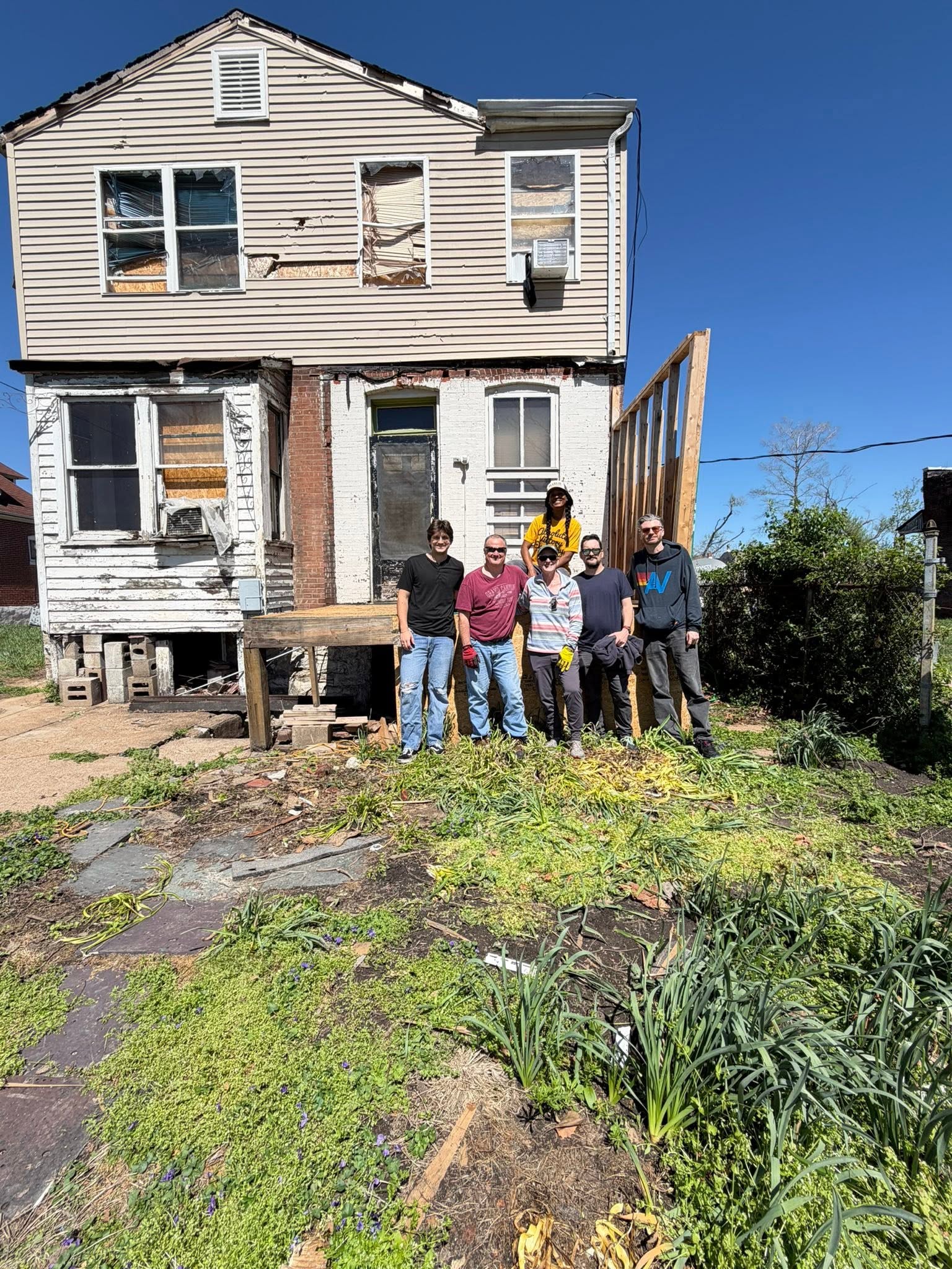 A group of six people posing together outdoors in front of a dilapidated, two-story house undergoing repairs.