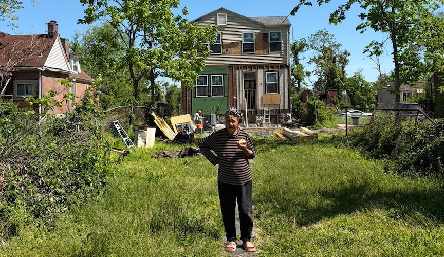 An older woman stands in an overgrown yard, taking a bite of food and looking towards the camera. Behind her, a two-story house is undergoing major exterior renovations with exposed framing and green sheathing.