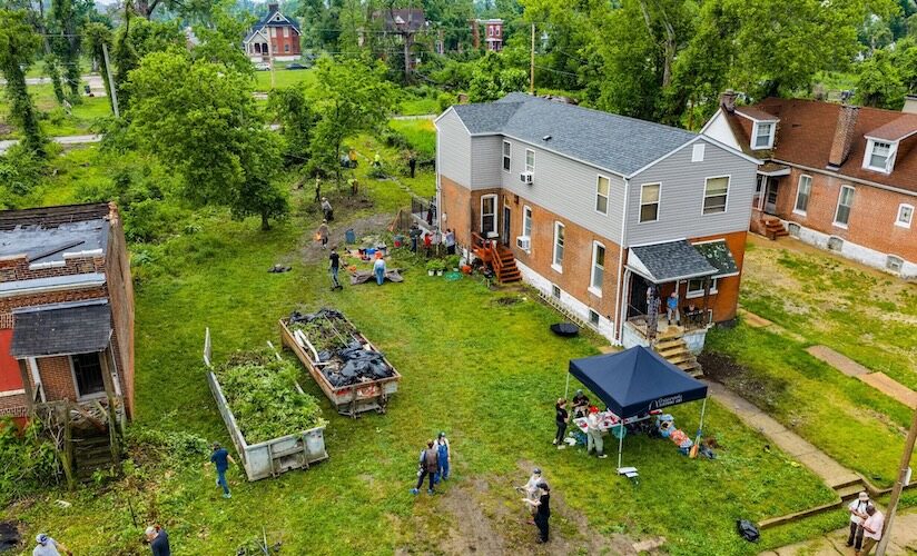 An aerial shot captures volunteers cleaning up a large yard next to a renovated two-story house, with a black "Grassroots Redeeming Love" tent set up on the lawn. Two large dumpsters on the grass are overflowing with cleared brush and yard waste.