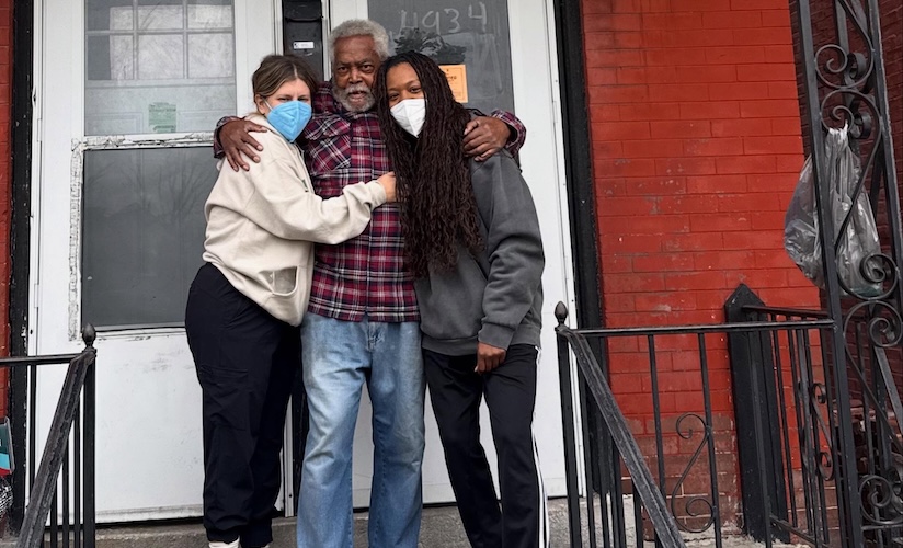Two younger women wearing face masks share a warm hug with an older man on the front steps of a red brick building. The trio smiles together in front of a white entry door.