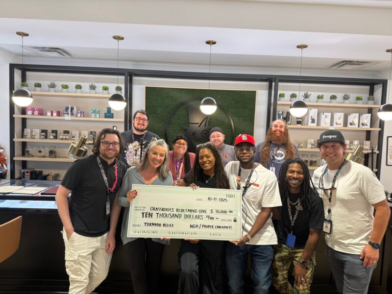 A diverse group of nine smiling people pose inside a retail store while holding a giant $10,000 donation check. The check is made out to Grassroots Redeeming Love for tornado relief from NGH and Proper Cannabis.