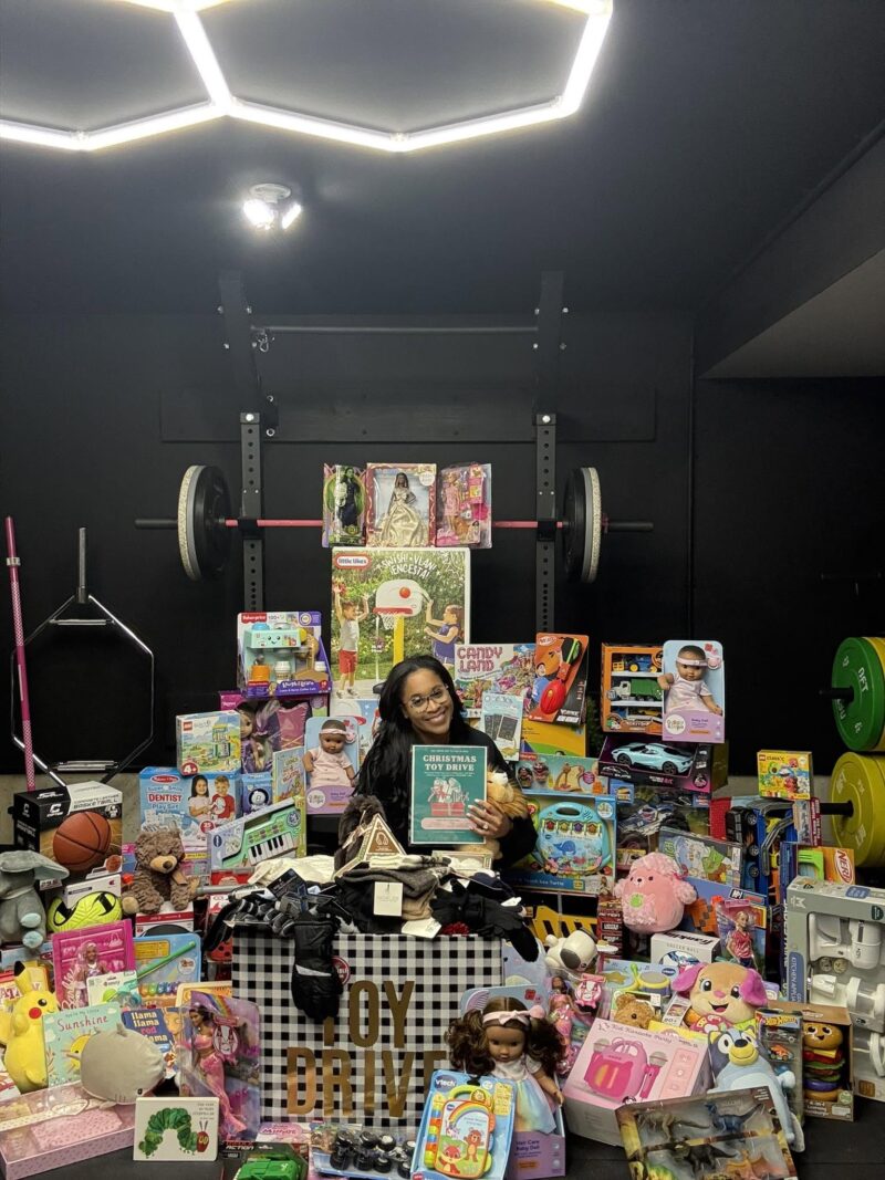 A smiling woman sits surrounded by a massive pile of donated toys, games, and gifts in a home gym. She is holding up a green flyer for a "Christmas Toy Drive."