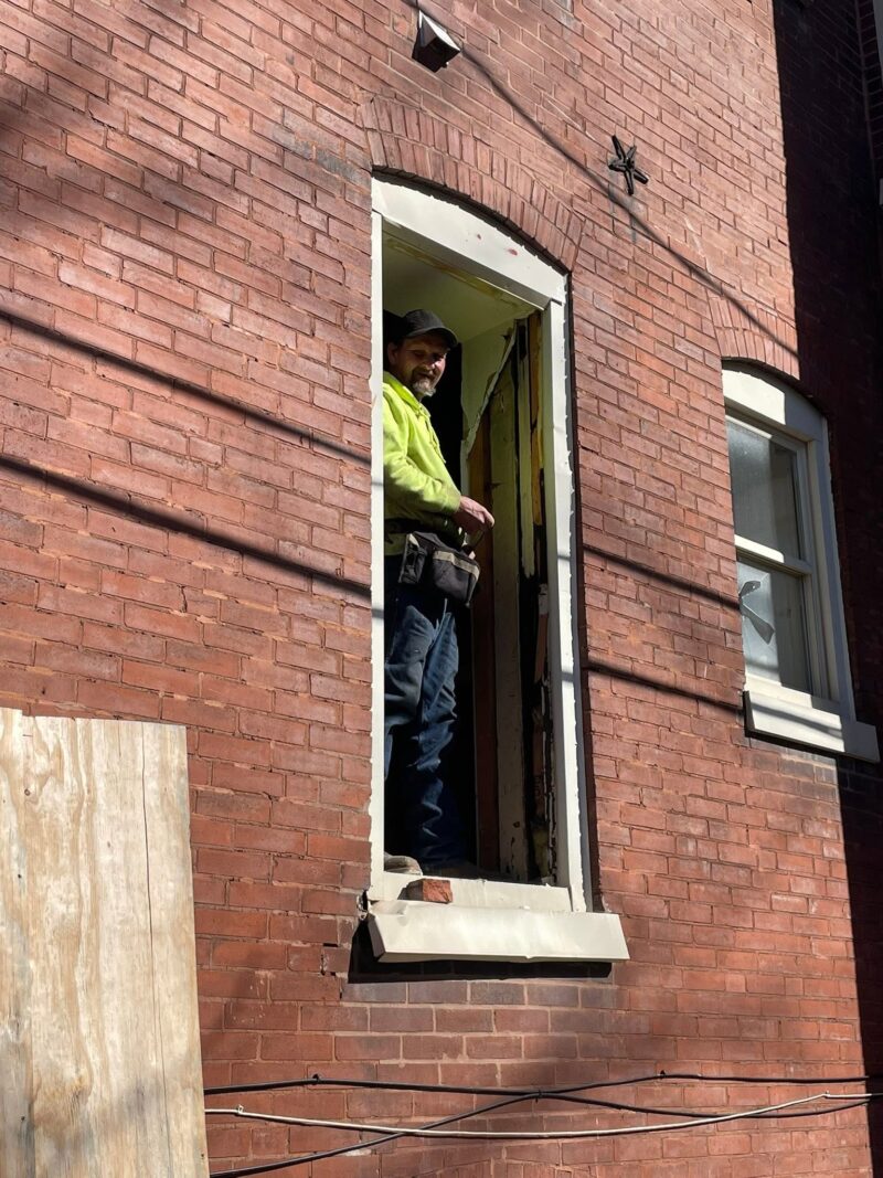 A smiling construction worker wearing a bright yellow shirt, a black cap, and a toolbelt stands inside the doorway of a red brick building looking outward.
