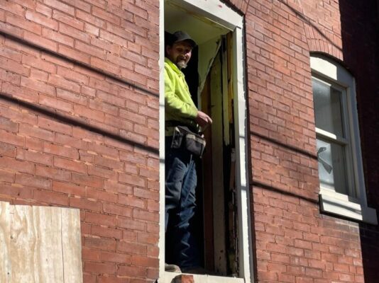 A smiling construction worker wearing a bright yellow shirt, a black cap, and a toolbelt stands inside the doorway of a red brick building looking outward.