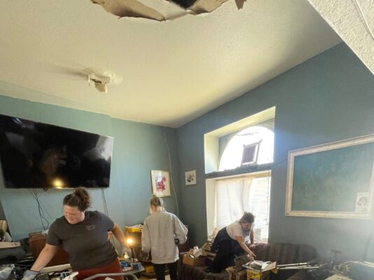 Three women work together to clear out a heavily cluttered room filled with random belongings, boxes, and luggage. The room features a blue wall and a significantly damaged ceiling.