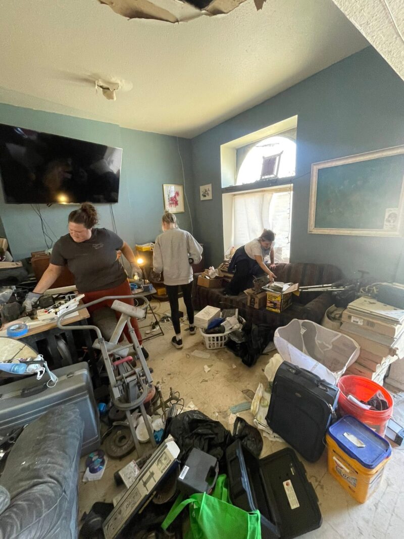 Three women work together to clear out a heavily cluttered room filled with random belongings, boxes, and luggage. The room features a blue wall and a significantly damaged ceiling.
