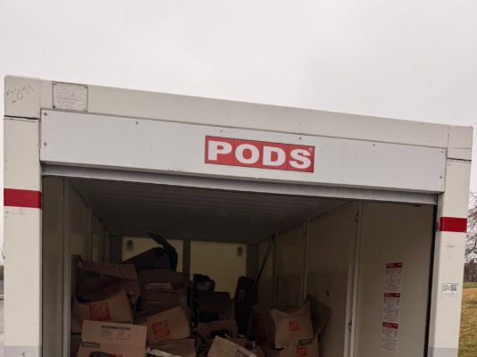 An open PODS moving container is packed tightly with cardboard Home Depot boxes, wicker hampers, and a laundry basket.
