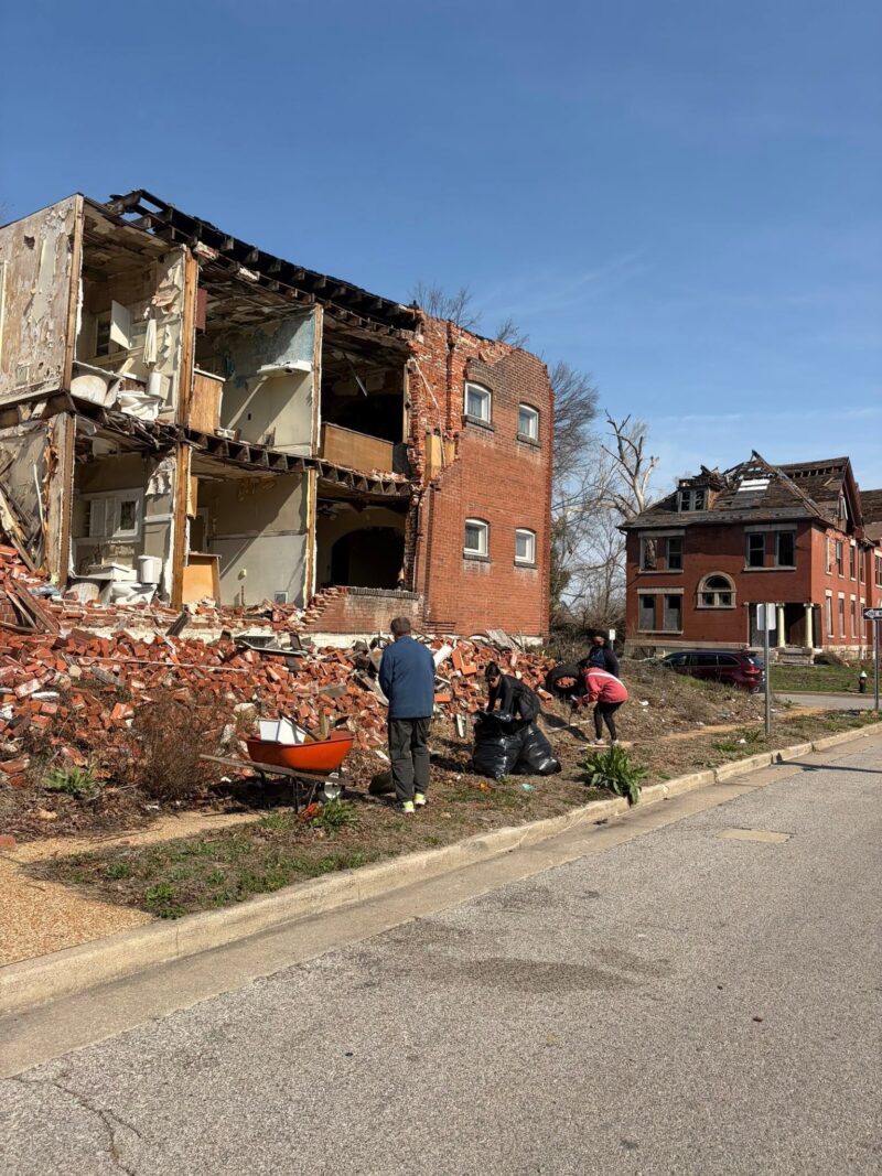 A group of volunteers cleans up the sidewalk in front of a multi-story brick building that has suffered a massive wall collapse. They are using a wheelbarrow and black trash bags to manage the debris next to a huge pile of fallen bricks.