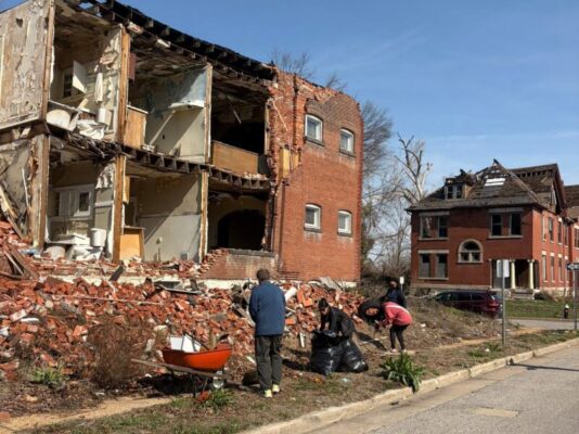 A group of volunteers cleans up the sidewalk in front of a multi-story brick building that has suffered a massive wall collapse. They are using a wheelbarrow and black trash bags to manage the debris next to a huge pile of fallen bricks.