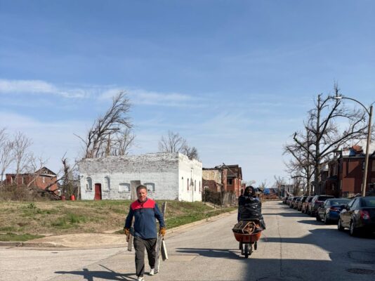 Two people haul debris down a residential street away from severely damaged brick buildings. One carries wooden boards while the other pushes a wheelbarrow full of trash bags.
