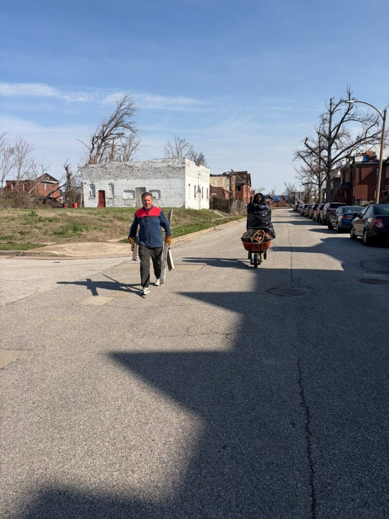 Two people haul debris down a residential street away from severely damaged brick buildings. One carries wooden boards while the other pushes a wheelbarrow full of trash bags.