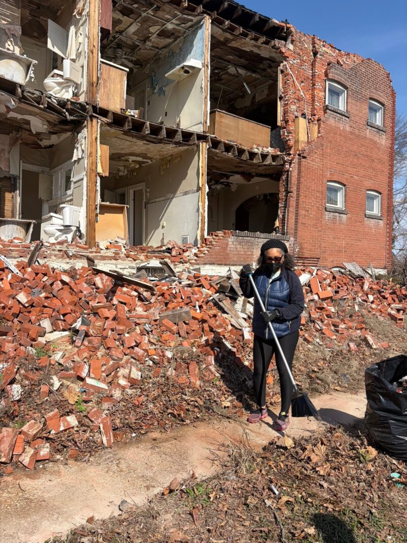 A woman wearing a face mask holds a broom while standing on a massive pile of red bricks from a collapsed multi-story building. The exposed interior of the ruined building, including a bathroom, is visible directly behind her.