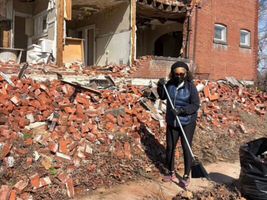 A woman wearing a face mask holds a broom while standing on a massive pile of red bricks from a collapsed multi-story building. The exposed interior of the ruined building, including a bathroom, is visible directly behind her.