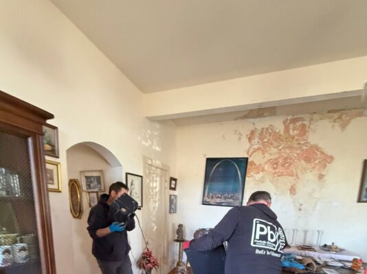 Two men carefully carry a large black object out of a messy room that features peeling wall paint and a red area rug.