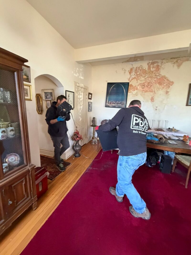 Two men carefully carry a large black object out of a messy room that features peeling wall paint and a red area rug.