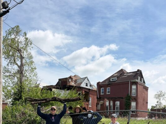 Four smiling volunteers stand behind a chain-link fence holding up a large, heavy fallen tree branch they just cleared. Behind them sit several large brick homes with severe roof and structural damage.