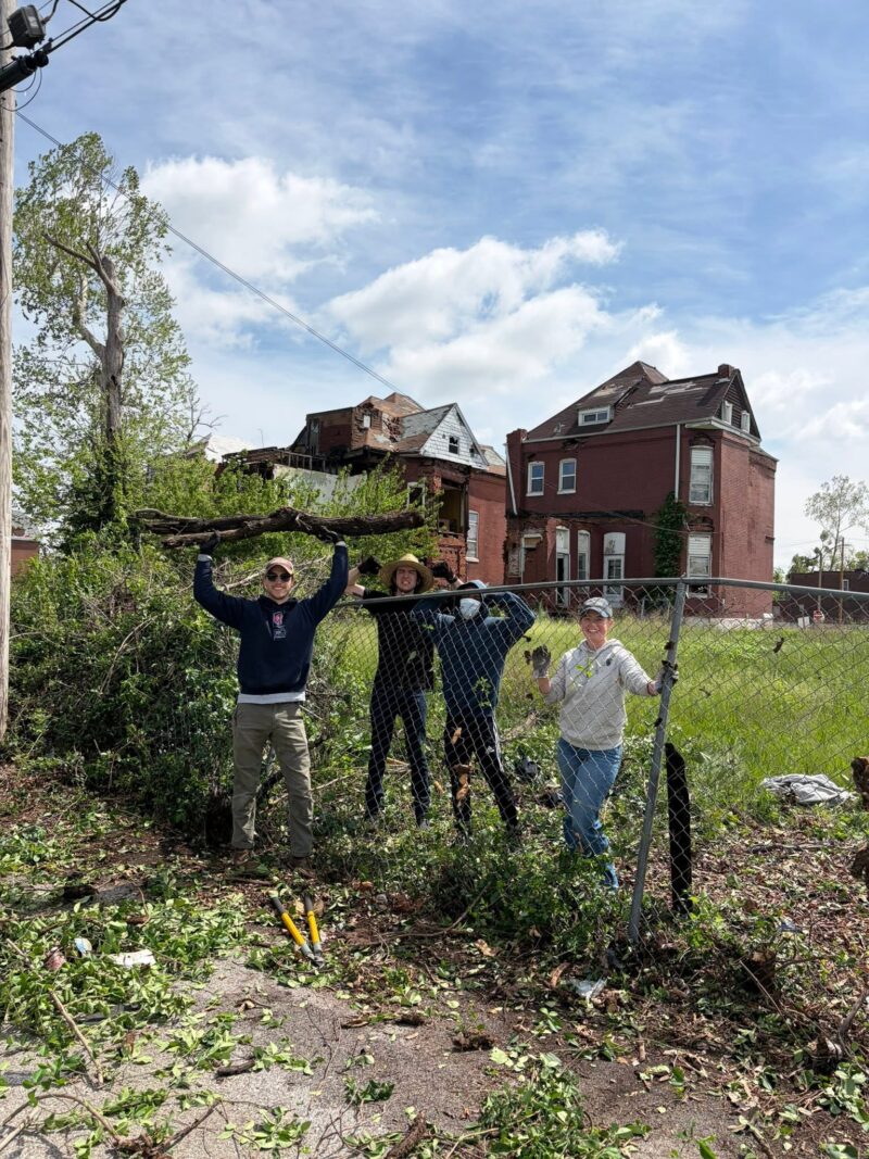 Four smiling volunteers stand behind a chain-link fence holding up a large, heavy fallen tree branch they just cleared. Behind them sit several large brick homes with severe roof and structural damage.
