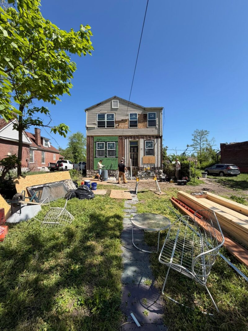 Construction workers are busy renovating the exterior of a two-story house, working around the exposed foundation and front entrance. The foreground is filled with construction debris, lawn furniture, and stacks of fresh lumber.