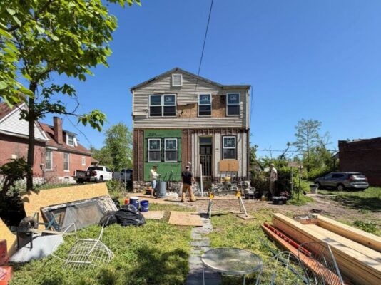 Construction workers are busy renovating the exterior of a two-story house, working around the exposed foundation and front entrance. The foreground is filled with construction debris, lawn furniture, and stacks of fresh lumber.