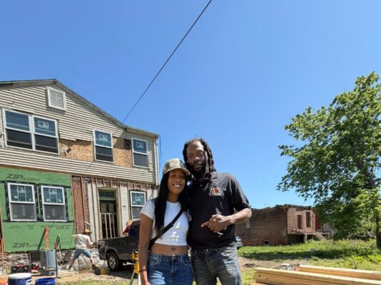 A smiling man and woman pose together in the yard of a house undergoing extensive renovations. The house behind them features exposed framing and green ZIP System sheathing panels on the lower level.