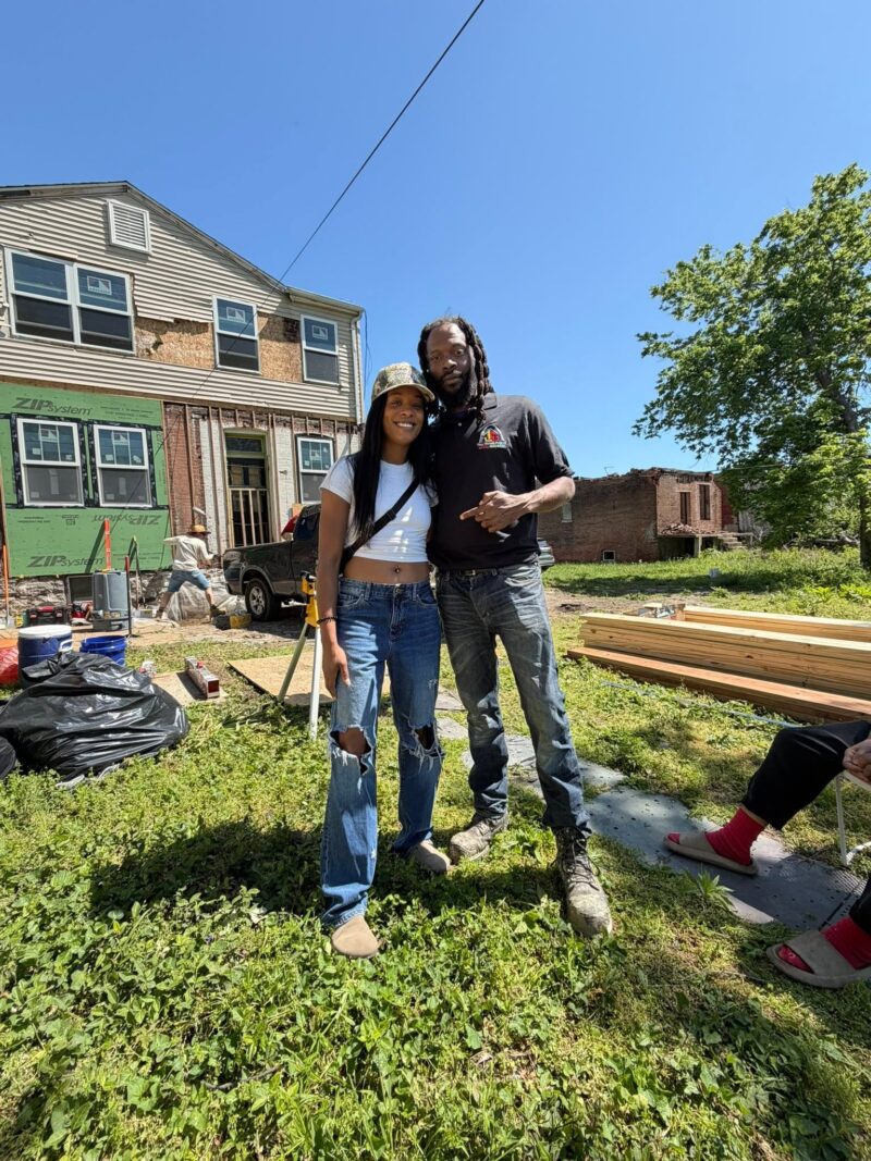A smiling man and woman pose together in the yard of a house undergoing extensive renovations. The house behind them features exposed framing and green ZIP System sheathing panels on the lower level.