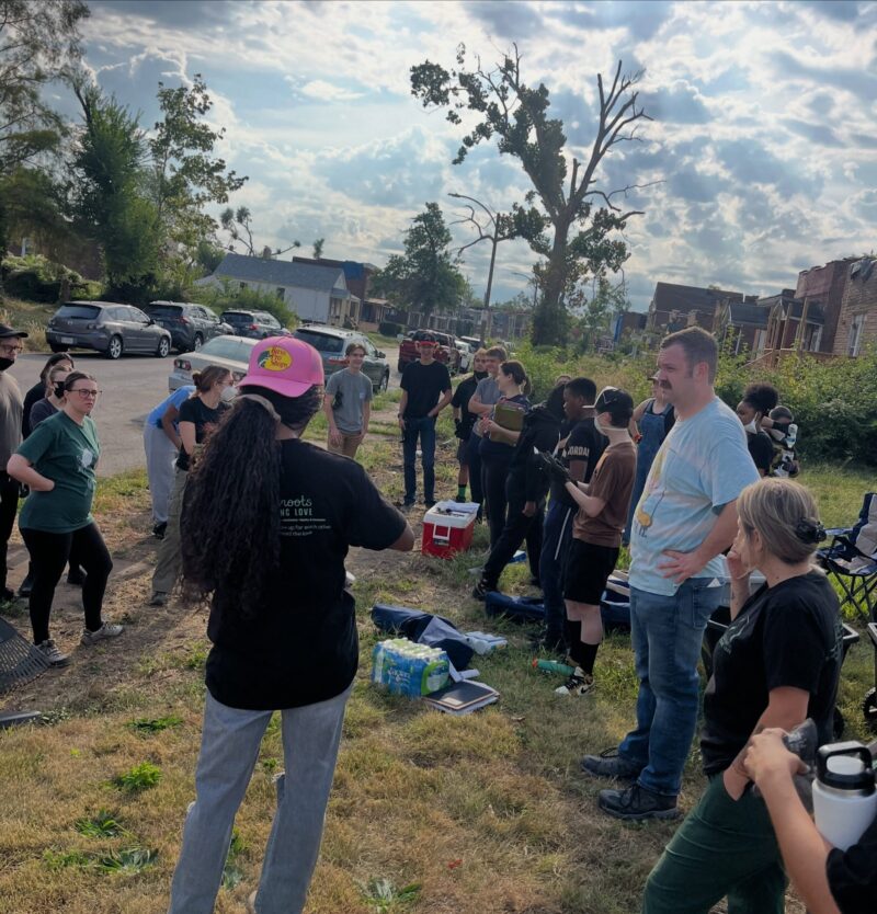 A woman wearing a pink hat stands facing a large group of volunteers gathered on a grassy curb alongside a residential street. She appears to be giving instructions or a briefing before a neighborhood cleanup event.